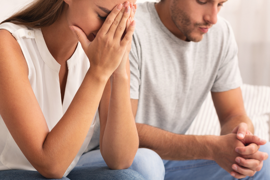 A couple sitting next to each other; the woman covers her face in distress while the man looks down with pursed lips, appearing guilty.